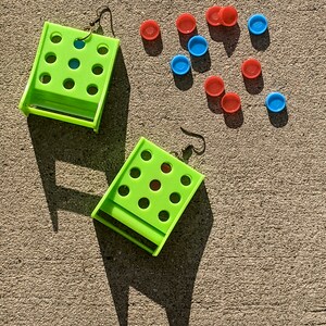 May include: A pair of lime green earrings with a grid of holes. The earrings are on a gray concrete surface with red and blue plastic circles scattered around them.