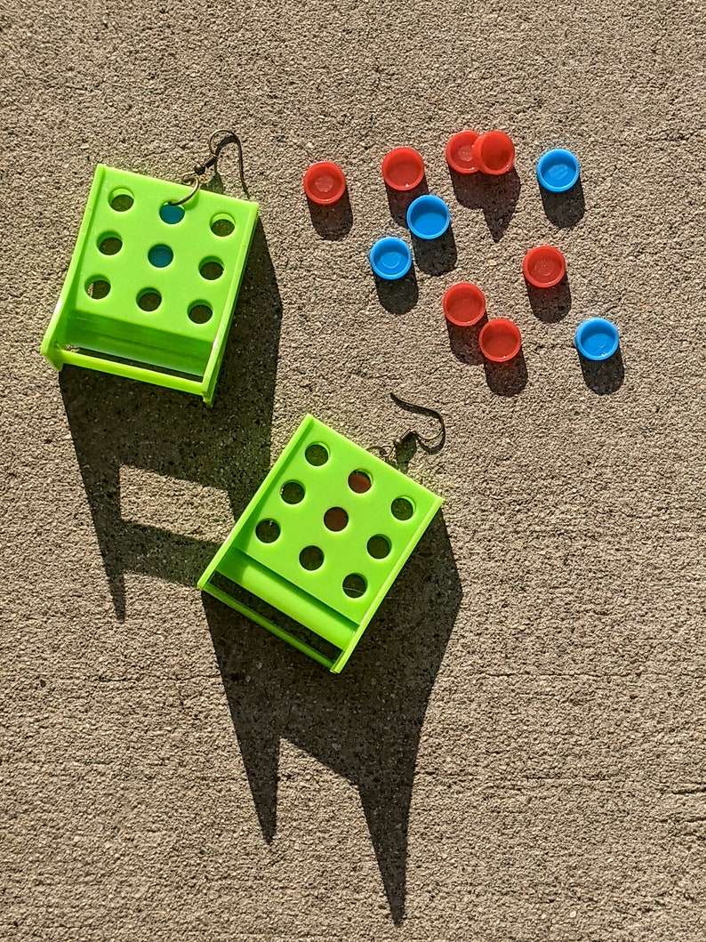 May include: A pair of lime green earrings with a grid of holes. The earrings are on a gray concrete surface with red and blue plastic circles scattered around them.