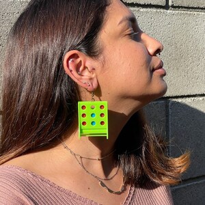 May include: A pair of green earrings shaped like a classic game of connect four. The earrings have red, blue, and green circles.