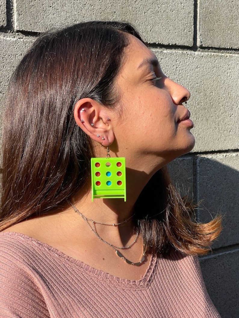 May include: A pair of green earrings shaped like a classic game of connect four. The earrings have red, blue, and green circles.