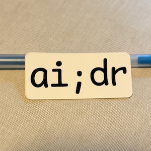 Puede incluir: Una tarjeta rectangular blanca con las letras "ai;dr" en fuente negra está colocada sobre un bolígrafo azul y transparente. La tarjeta está centrada y el bolígrafo corre horizontalmente detrás de ella. El fondo es una tela beige claro.