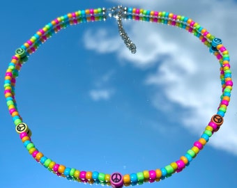 Puede incluir: Un collar de cuentas de colores con dijes de signo de paz. El collar está hecho con cuentas de color rosa brillante, verde, amarillo y azul.