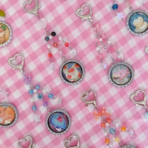 May include: Keychains featuring colorful cartoon character designs within bottle cap frames. Each keychain has a heart-shaped clasp and is adorned with beaded accents in various colors, including pink, blue, and orange. The background is a pink and white checkered pattern.