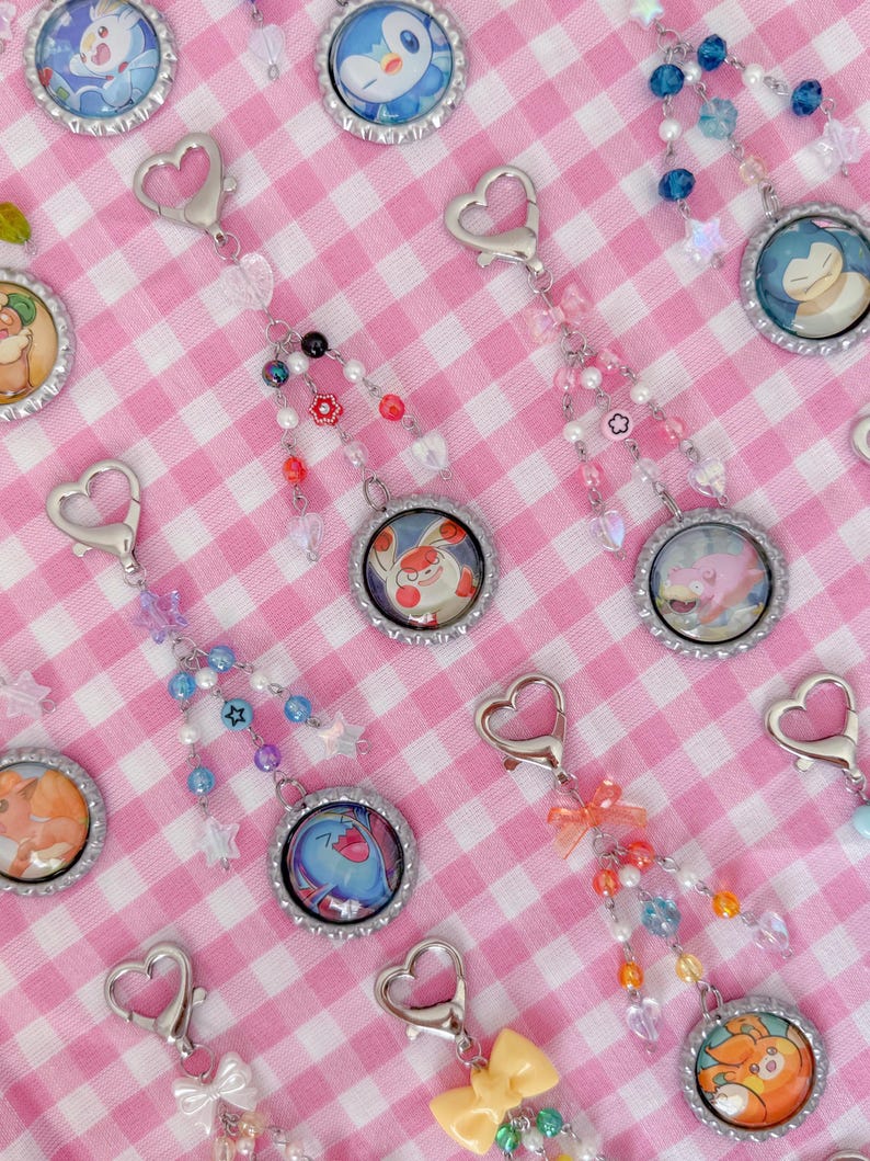 May include: Keychains featuring colorful cartoon character designs within bottle cap frames. Each keychain has a heart-shaped clasp and is adorned with beaded accents in various colors, including pink, blue, and orange. The background is a pink and white checkered pattern.