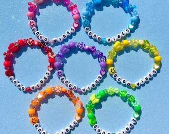 May include: Six colorful beaded bracelets with the words "Good Vibes" spelled out on each one. The bracelets are made with different colored beads, including smiley faces, hearts, butterflies, and flowers.