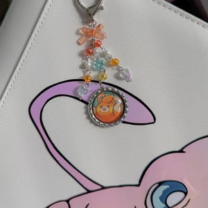 May include: A silver charm with a heart-shaped clasp, a small orange bow, and colorful beads. The charm features a circular pendant with an orange and yellow cartoon character inside a silver frame. The charm is attached to a white surface with a pink and purple cartoon graphic.