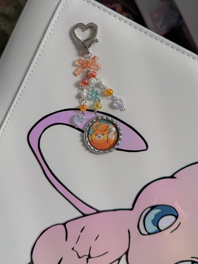 May include: A silver charm with a heart-shaped clasp, a small orange bow, and colorful beads. The charm features a circular pendant with an orange and yellow cartoon character inside a silver frame. The charm is attached to a white surface with a pink and purple cartoon graphic.