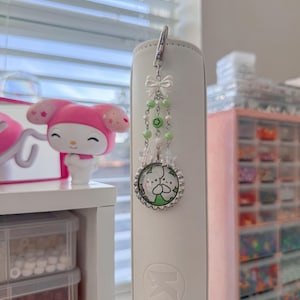 May include: A white and silver bookmark charm with a cartoon character design. The charm features a silver clasp, a white bow, green beads, and a circular pendant with a cartoon character. The charm is attached to a white book.