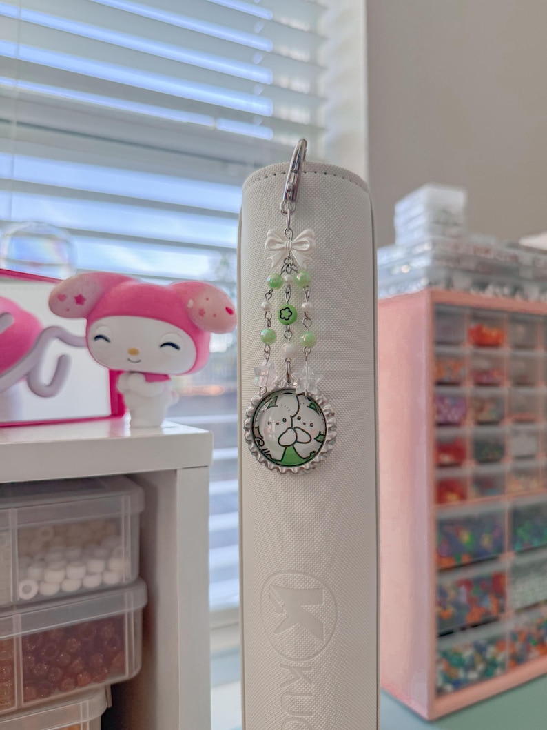 May include: A white and silver bookmark charm with a cartoon character design. The charm features a silver clasp, a white bow, green beads, and a circular pendant with a cartoon character. The charm is attached to a white book.