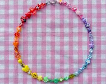 May include: A rainbow-colored beaded necklace featuring a variety of shapes including stars, flowers, hearts, a ladybug, a smiley face, and a green die. The necklace is arranged in a spectrum of colors and includes a silver-toned metal clasp.