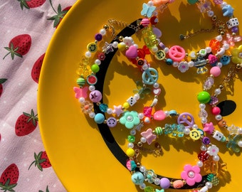May include: Colorful beaded necklaces with various charms, including flowers, butterflies, smiley faces, and peace signs, are displayed on a yellow plate. The necklaces feature a mix of pastel and bright beads. A pink strawberry-patterned fabric is in the background.