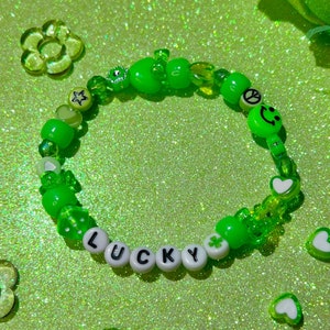 Könnte beinhalten: Grünes und weißes Perlenarmband mit dem Wort "LUCKY" in weißen Perlen. Das Armband enthält auch grüne Herz-, Stern- und Friedenszeichenperlen.