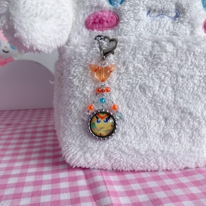 May include: A silver-toned keychain with a heart-shaped clasp and a charm featuring a cartoon owl face. The charm is adorned with orange, blue, and clear beads. The keychain is attached to a white plush toy with blue and pink accents.