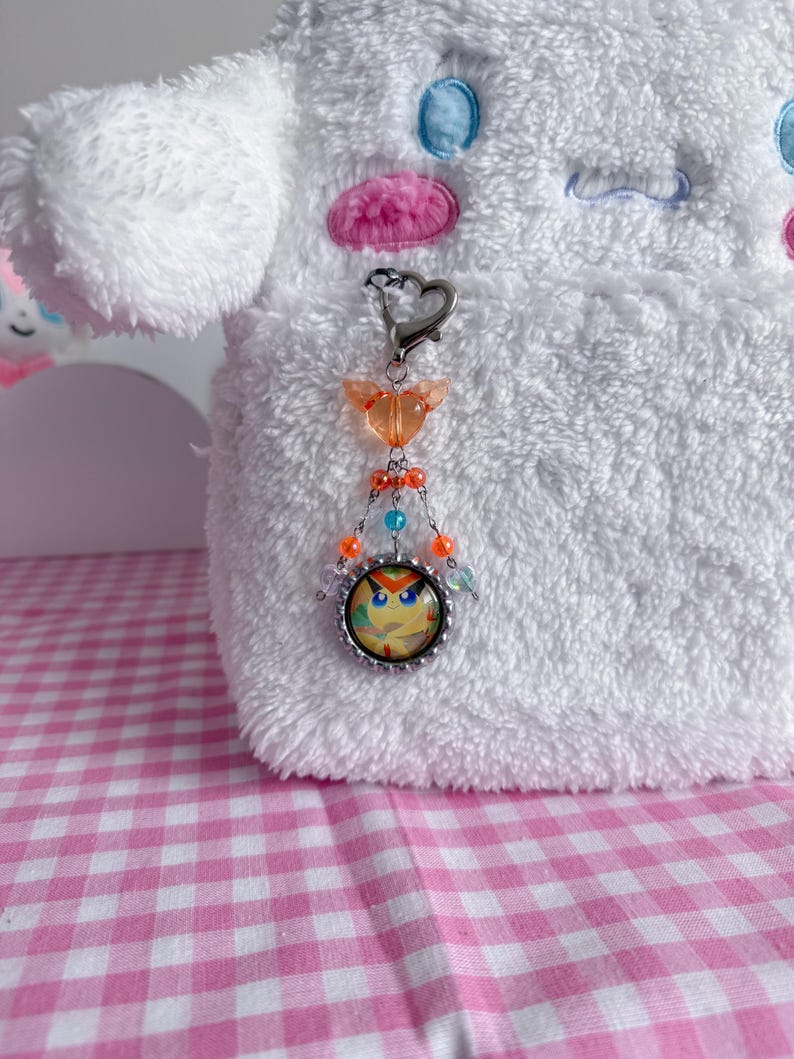 May include: A silver-toned keychain with a heart-shaped clasp and a charm featuring a cartoon owl face. The charm is adorned with orange, blue, and clear beads. The keychain is attached to a white plush toy with blue and pink accents.