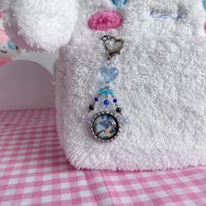 May include: A silver-toned keychain with a heart-shaped clasp and a charm featuring a cartoon image. The charm is adorned with blue and clear beads. The background is a white plush toy and a pink and white checkered surface.