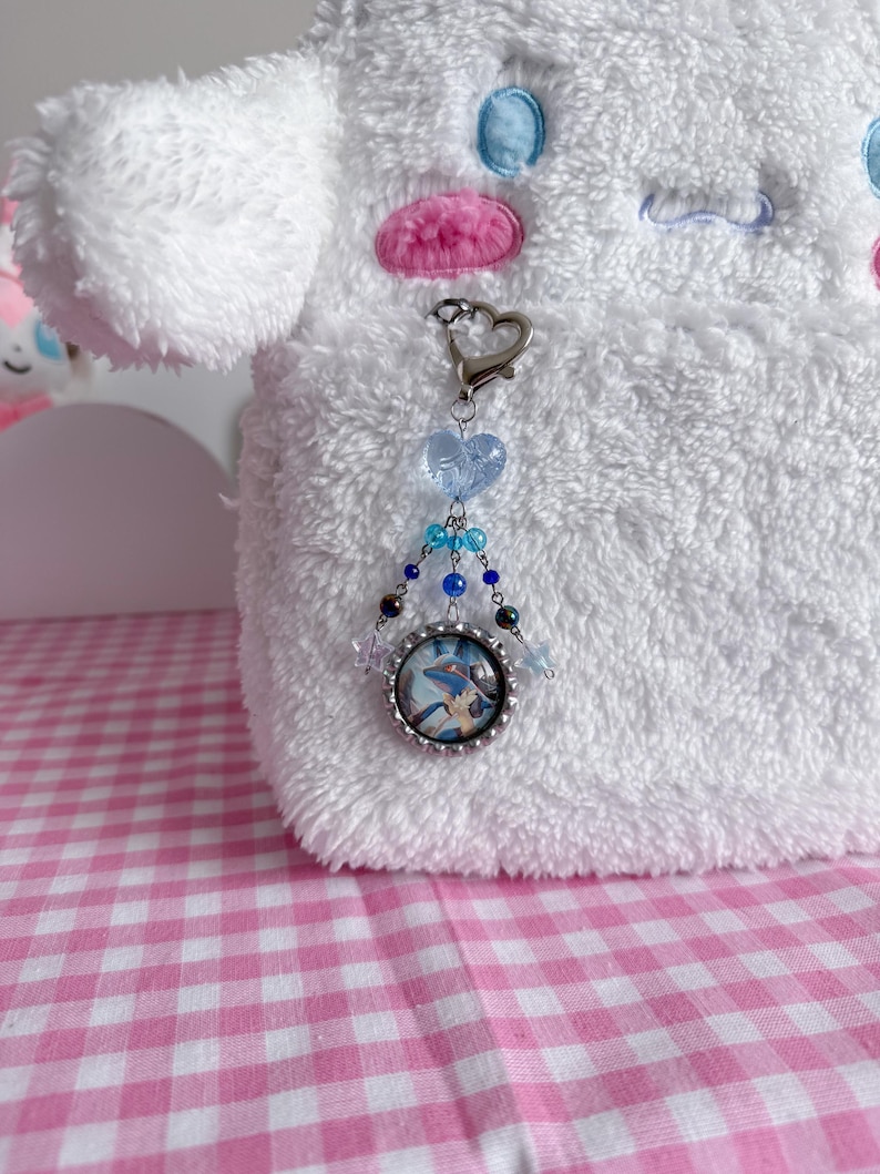 May include: A silver-toned keychain with a heart-shaped clasp and a charm featuring a cartoon image. The charm is adorned with blue and clear beads. The background is a white plush toy and a pink and white checkered surface.