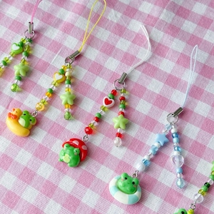 May include: Four phone charms featuring green frog figures. Each charm has a colorful beaded strand with star, heart, or round beads. The frogs are depicted with a yellow crescent moon, a red mushroom cap, a blue and white life ring, and a plain green frog.
