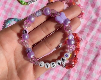 May include: A purple and clear beaded bracelet with the name "LYSSA" spelled out in white beads. The bracelet also features a purple butterfly charm, a peace sign charm, a star charm, and a heart charm.