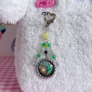 May include: A beaded keychain with a frog design. The keychain features a silver-colored heart-shaped clasp, yellow and green beads, and a circular pendant with a frog illustration in a bottle cap.