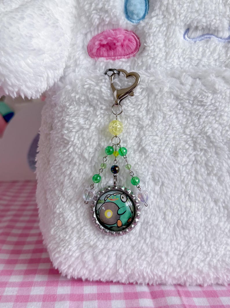 May include: A beaded keychain with a frog design. The keychain features a silver-colored heart-shaped clasp, yellow and green beads, and a circular pendant with a frog illustration in a bottle cap.