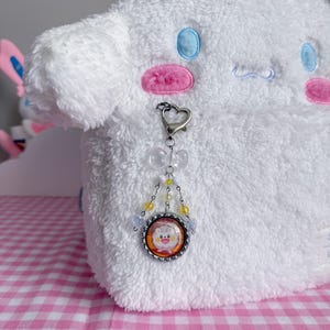 May include: A white plush toy with blue and pink accents, featuring a heart-shaped clasp and a charm with a cartoon character image. The charm is adorned with yellow and clear beads.