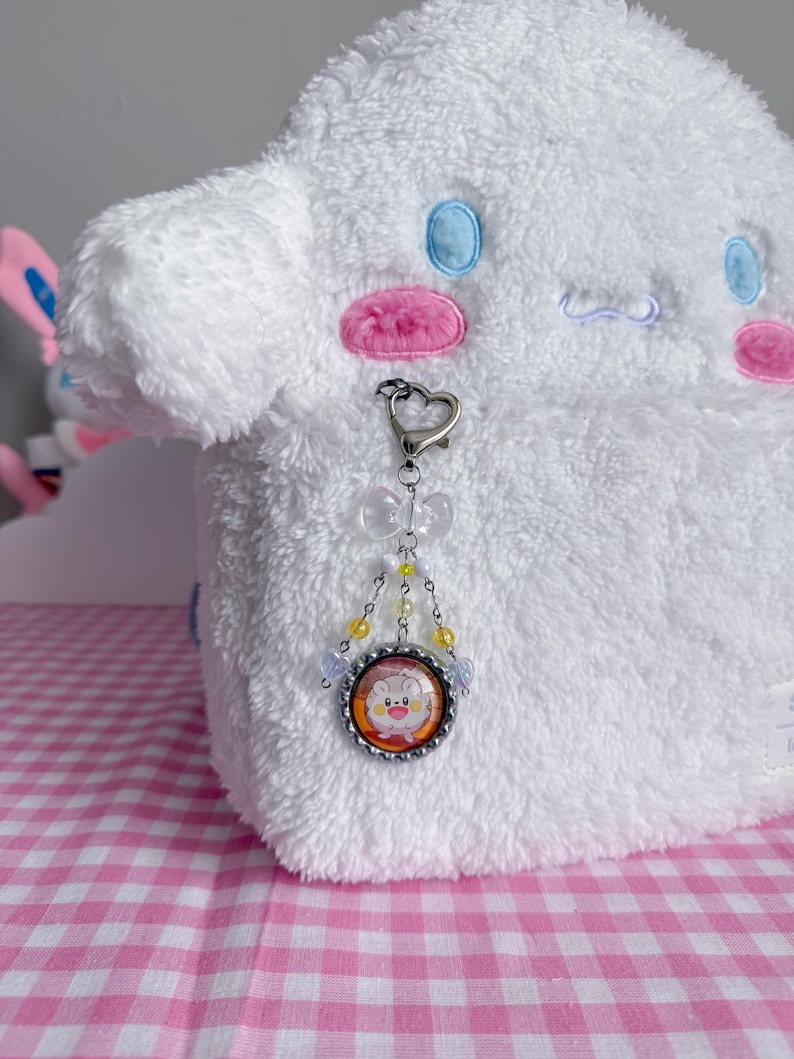May include: A white plush toy with blue and pink accents, featuring a heart-shaped clasp and a charm with a cartoon character image. The charm is adorned with yellow and clear beads.