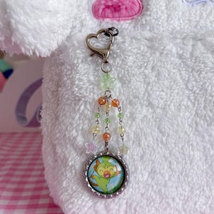 May include: A silver-toned keychain with a heart-shaped clasp and a charm featuring a cartoon character. The charm is surrounded by a beaded chain with green, orange, and clear beads, and star-shaped accents. The background is a white, fluffy material.