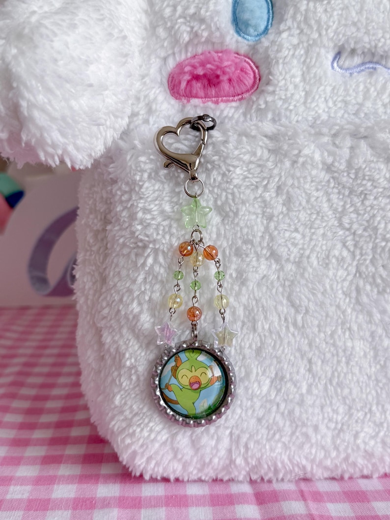 May include: A silver-toned keychain with a heart-shaped clasp and a charm featuring a cartoon character. The charm is surrounded by a beaded chain with green, orange, and clear beads, and star-shaped accents. The background is a white, fluffy material.