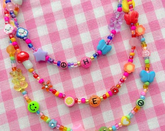 May include: Three colorful beaded necklaces with various charms, including hearts, stars, mushrooms, butterflies, and peace signs. The necklaces spell out the word "HER" in colorful letters.