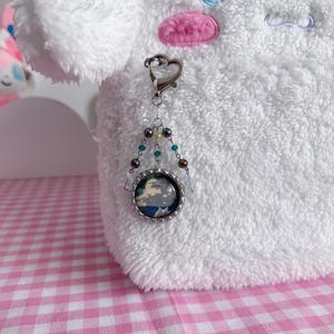 May include: A silver keychain with a heart-shaped clasp and a beaded charm. The charm features a small, round image with a blue and white design, possibly a landscape or abstract art. The background is a white, fluffy plush toy.