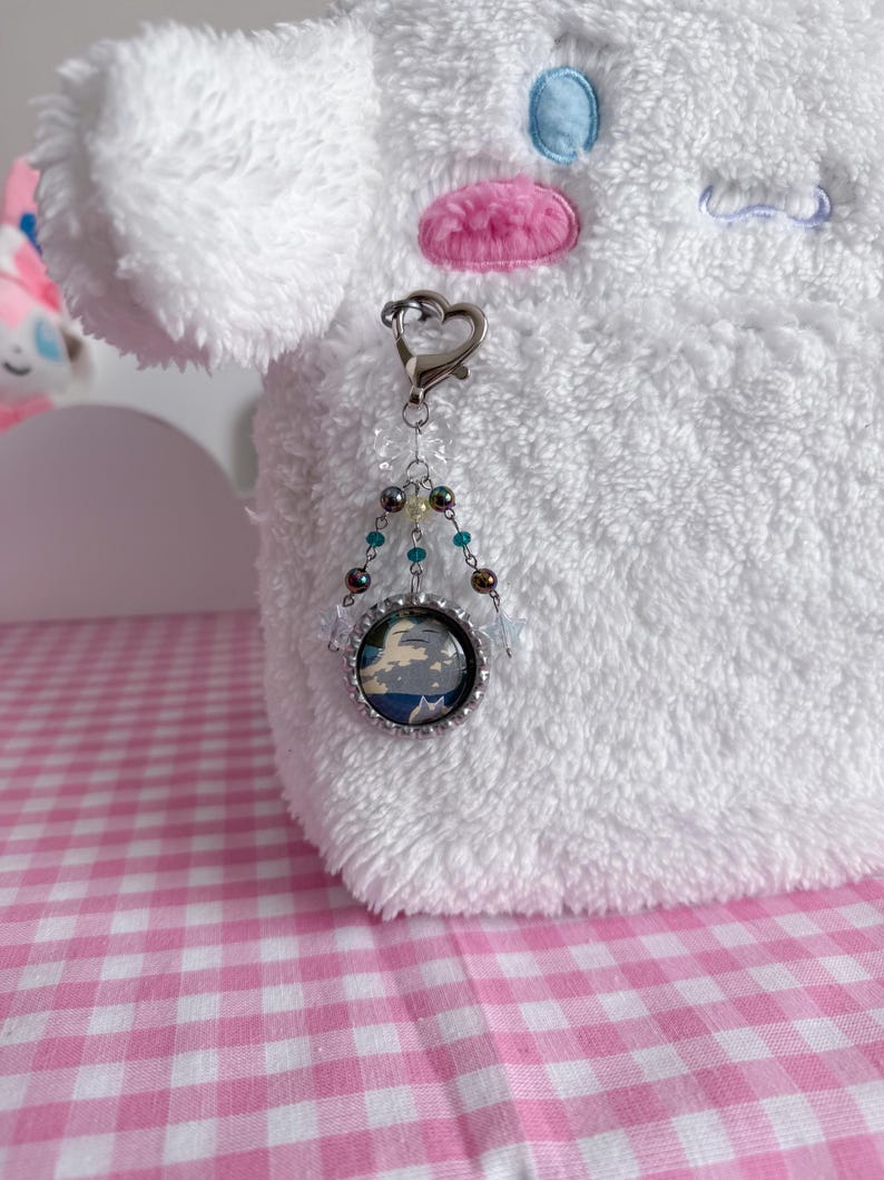 May include: A silver keychain with a heart-shaped clasp and a beaded charm. The charm features a small, round image with a blue and white design, possibly a landscape or abstract art. The background is a white, fluffy plush toy.