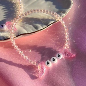 May include: A pink crystal beaded necklace with pink angel wing charms and white beads with the number "4" on them. The necklace is on a pink surface.
