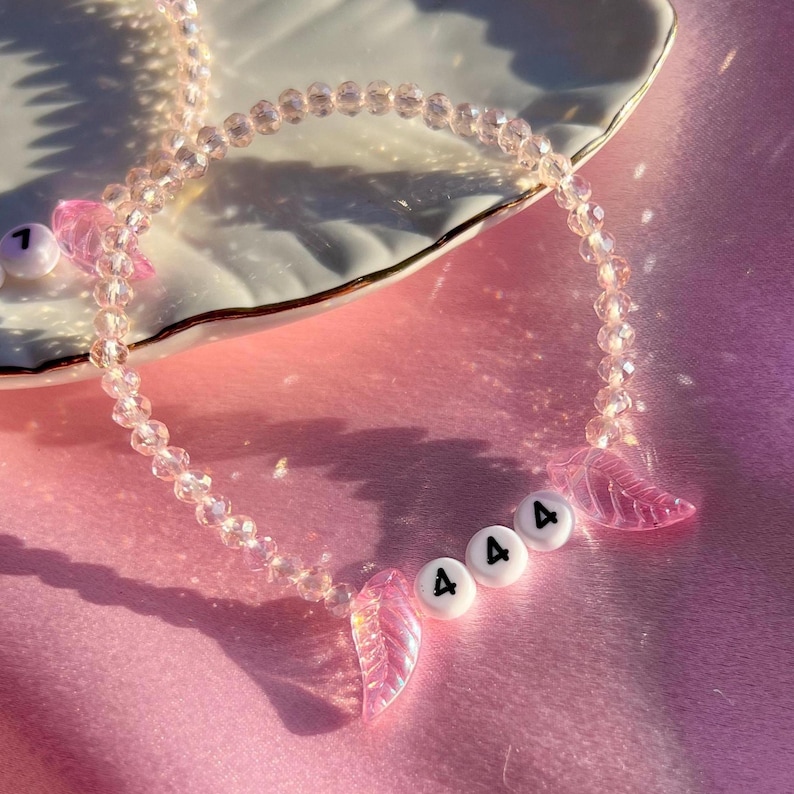 May include: A pink crystal beaded necklace with pink angel wing charms and white beads with the number "4" on them. The necklace is on a pink surface.