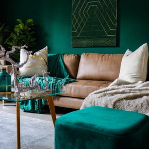 May include: A teal green velvet ottoman with gold legs sits in front of a brown leather sofa with a white throw blanket. The sofa is in a living room with a green wall and a gold geometric print on the wall.