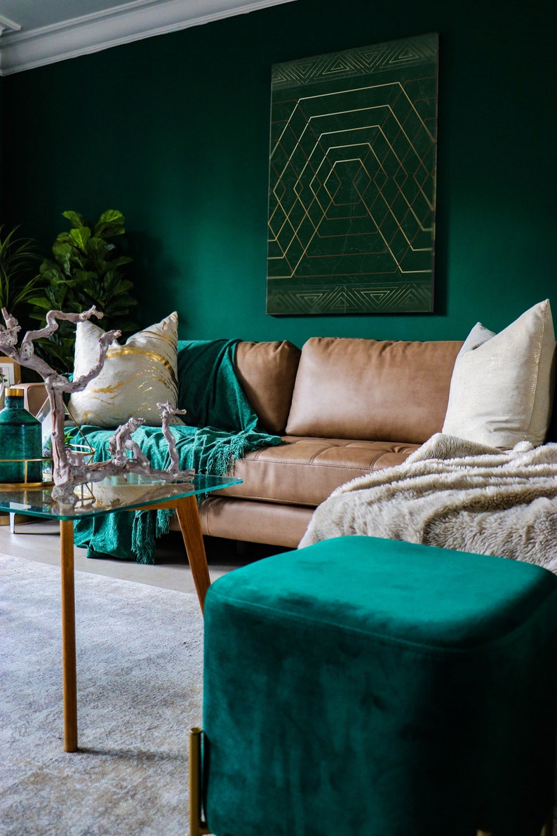 May include: A teal green velvet ottoman with gold legs sits in front of a brown leather sofa with a white throw blanket. The sofa is in a living room with a green wall and a gold geometric print on the wall.
