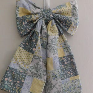 May include: A large, decorative fabric bow with a patchwork design. The bow features squares of floral and geometric patterns in shades of gray, yellow, and blue. It is hanging from a white hook on a white wall, suggesting a home decor item.