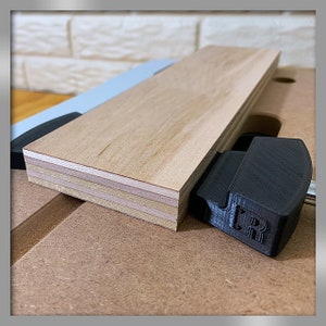 Könnte beinhalten: Nahaufnahme eines Holzstücks, das auf einer Werkbank befestigt ist. Das Holz ist hellbraun und hat eine mehrschichtige Konstruktion. Die Klemme ist schwarz und hat eine strukturierte Oberfläche. Die Klemme ist mit "TR" beschriftet.