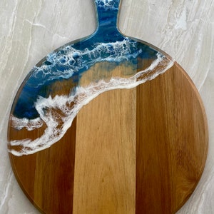 May include: Round wooden cutting board with a blue and white resin ocean wave design. The board has a handle and is perfect for serving cheese, charcuterie, or other appetizers.