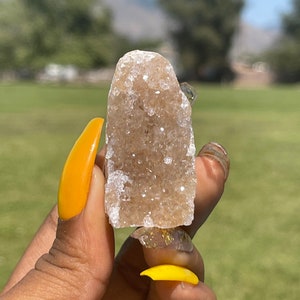 May include: A light brown crystal cluster with a sparkling, textured surface. The crystal is held in a hand with bright yellow nail polish. The background is a blurred green field and trees.