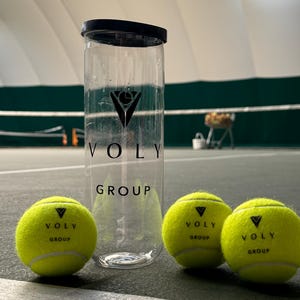 May include: A clear plastic container with a black lid and the "VOLY GROUP" logo, next to three yellow tennis balls. The tennis balls also have the "VOLY GROUP" logo. The background shows a tennis court.