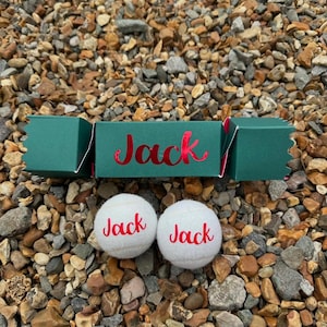 Op de afbeelding: Een donkergroene feestkraker met de naam "Jack" in rood script, samen met twee witte tennisballen, elk met "Jack" in het rood gedrukt. De items zijn gerangschikt op een bed van kleine, veelkleurige kiezelstenen.
