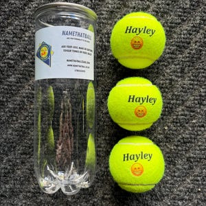 Könnte beinhalten: Drei gelbe Tennisbälle mit dem Namen "Hayley" und einem Smiley neben einem transparenten Plastikrohr mit der Aufschrift "NAMETHATBALL". Das Rohr enthält Kontaktinformationen.