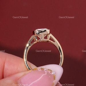 May include: A gold ring with a square-cut diamond. The ring has a delicate band and a bezel setting that holds the diamond securely. The ring is held up against a red background.
