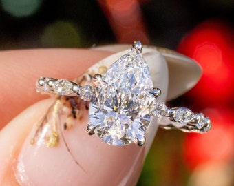 Könnte beinhalten: Ein funkelnder Verlobungsring mit einem birnenförmigen Diamanten als Mittelpunkt. Der Ring hat ein zartes Band, das mit kleineren, runden Diamanten besetzt ist. Der Ring wird vor einem verschwommenen Hintergrund aus Rot und Grün gehalten.