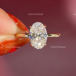 May include: A gold engagement ring featuring a large, oval-cut diamond. The diamond is the focal point, set on a delicate gold band. The ring is held against a blurred, reddish-purple background.