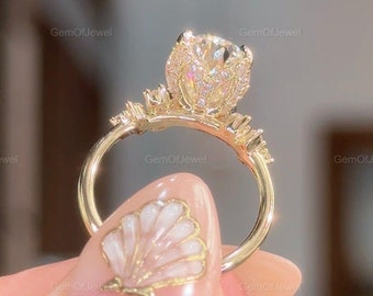 Puede incluir: Un anillo de oro con una gran piedra preciosa redonda y transparente engastada en un diseño con forma de flor. La banda del anillo está adornada con piedras preciosas más pequeñas. El anillo está en un dedo con un diseño de uña en forma de concha.