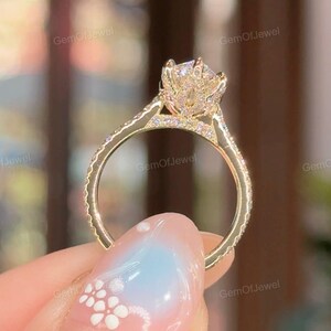 Puede incluir: Un anillo de compromiso de oro con una piedra central grande y brillante y piedras de acento más pequeñas a lo largo de la banda. El anillo tiene un diseño floral alrededor de la piedra central. El anillo es sostenido por una mano con un diseño de uñas azul claro y blanco.