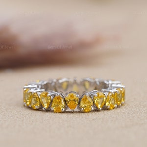 May include: A pear-shaped yellow gemstone eternity band ring set in white metal. The ring is made up of a row of yellow gemstones.