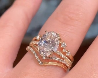 May include: A gold engagement ring with a large oval-shaped diamond center stone and smaller diamonds set in a delicate band. The ring is worn on a finger with a gold band on the finger next to it.