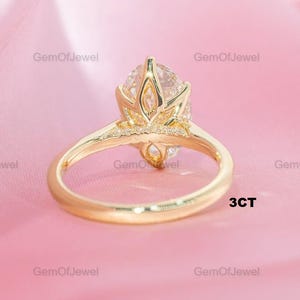 Pode incluir: Um anel de noivado dourado com um diamante de corte oval. O anel tem uma banda delicada e uma cravação detalhada. O texto "3CT" é visível. O fundo é rosa suave.